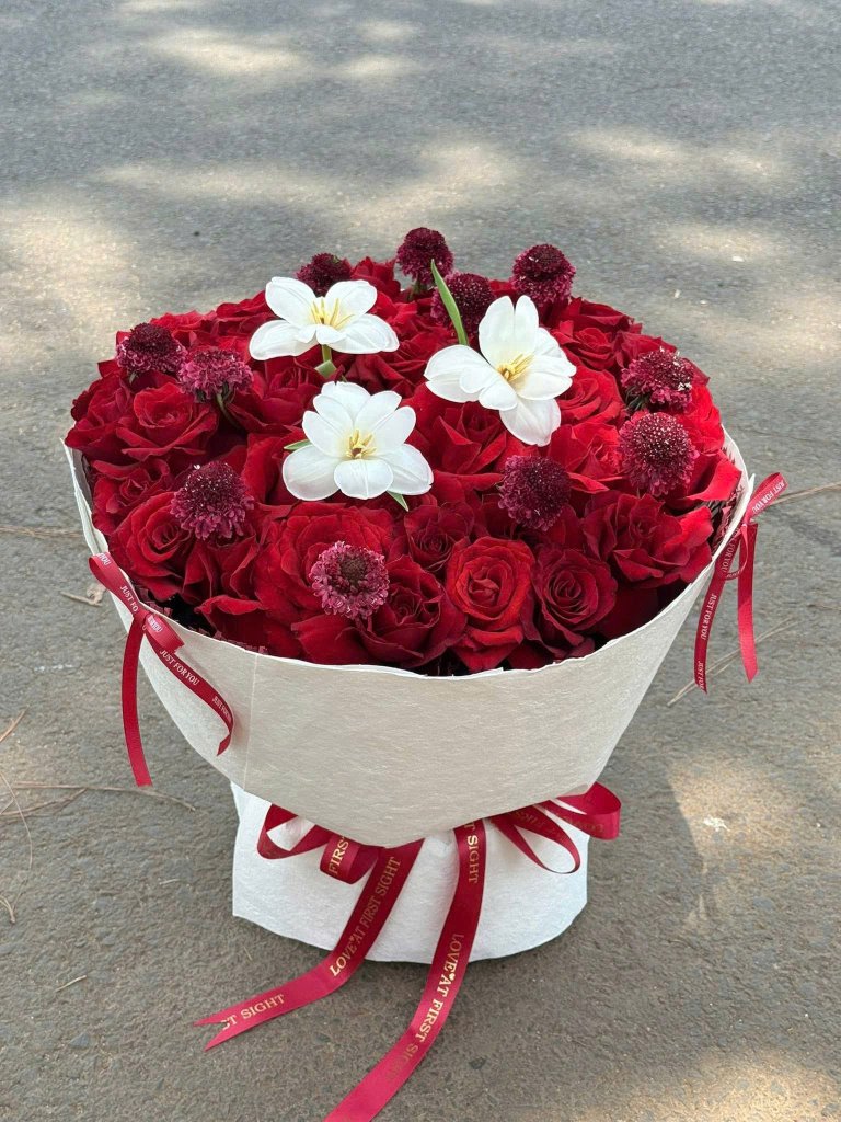 Bó hoa hồng đỏ tươi thắm kết hợp hoa trắng và phụ liệu trang trí, với ruy băng "JUST FOR YOU" và "LOVE AT FIRST SIGHT" từ ST