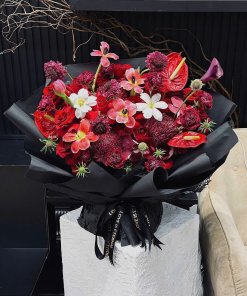Bó hoa đỏ rực rỡ với hồng nhung, loa kèn đỏ, cẩm chướng được gói tinh tế trong giấy đen, thắt nơ "LOVE IS ETERNAL" tại STflo