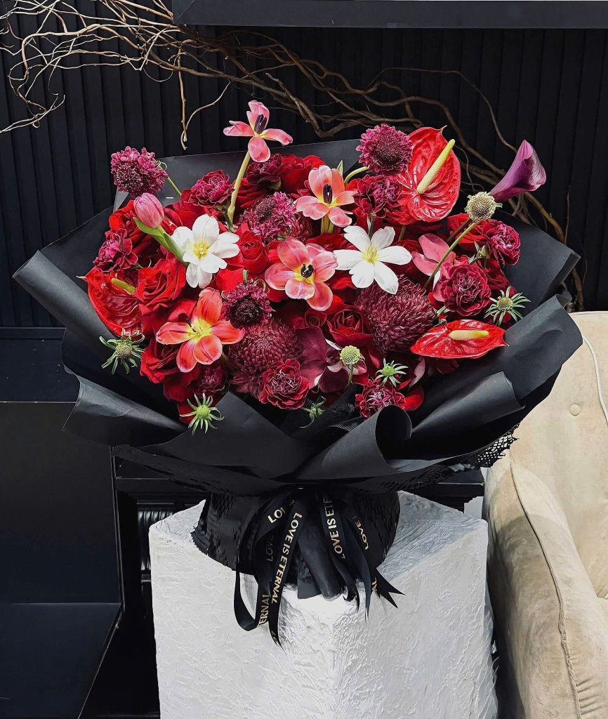 Bó hoa đỏ rực rỡ với hồng nhung, loa kèn đỏ, cẩm chướng được gói tinh tế trong giấy đen, thắt nơ "LOVE IS ETERNAL" tại STflo