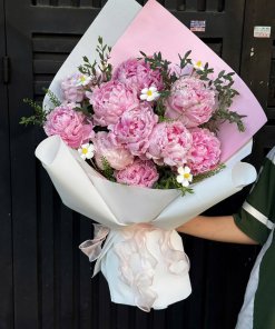 Bó hoa mẫu đơn hồng với những bông hoa tròn đầy, cánh hoa mềm mại, được gói trong giấy trắng và hồng, điểm xuyết lá xanh, tạ