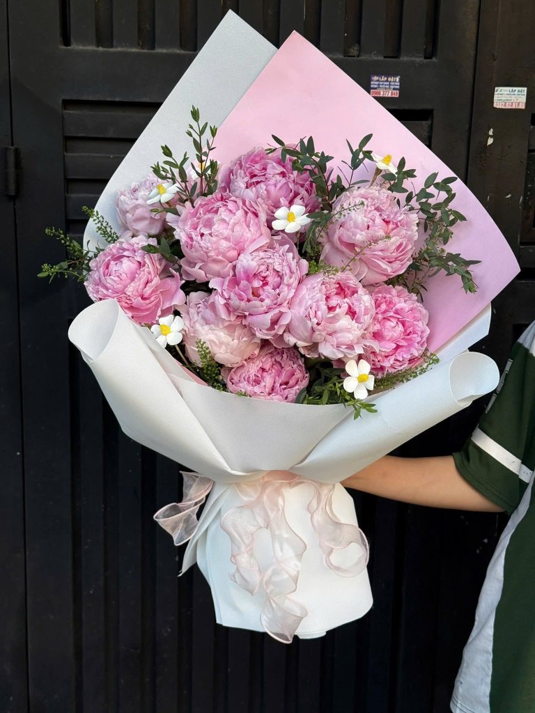 Bó hoa mẫu đơn hồng với những bông hoa tròn đầy, cánh hoa mềm mại, được gói trong giấy trắng và hồng, điểm xuyết lá xanh, tạ