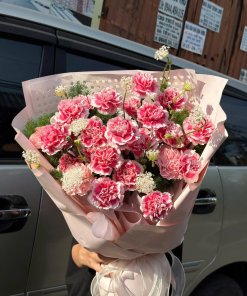 Bó hoa cẩm chướng hồng thắm rực rỡ được gói bằng giấy hồng và nơ trắng, khách hàng cầm bó hoa trước xe ô tô, STflowers