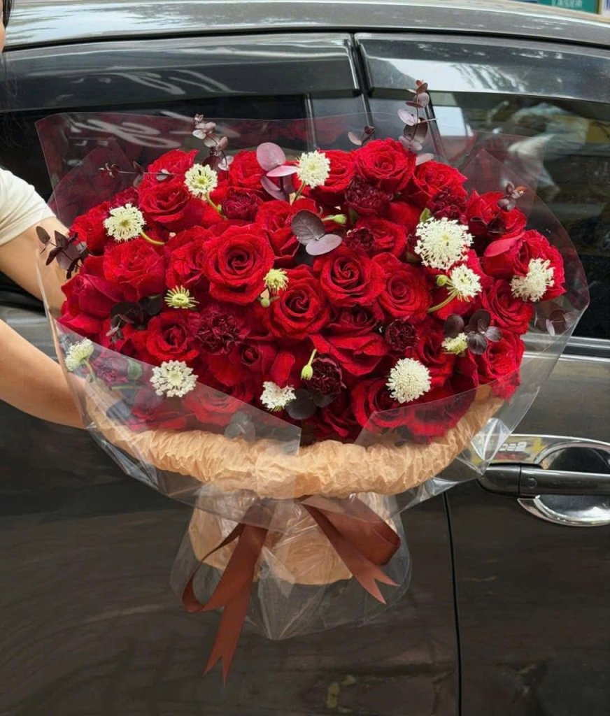 Bó hoa hồng đỏ rực rỡ sang trọng được gói cẩn thận, điểm xuyết hoa trắng nhỏ và lá phụ, thể hiện tình yêu nồng cháy. Đặt hoa
