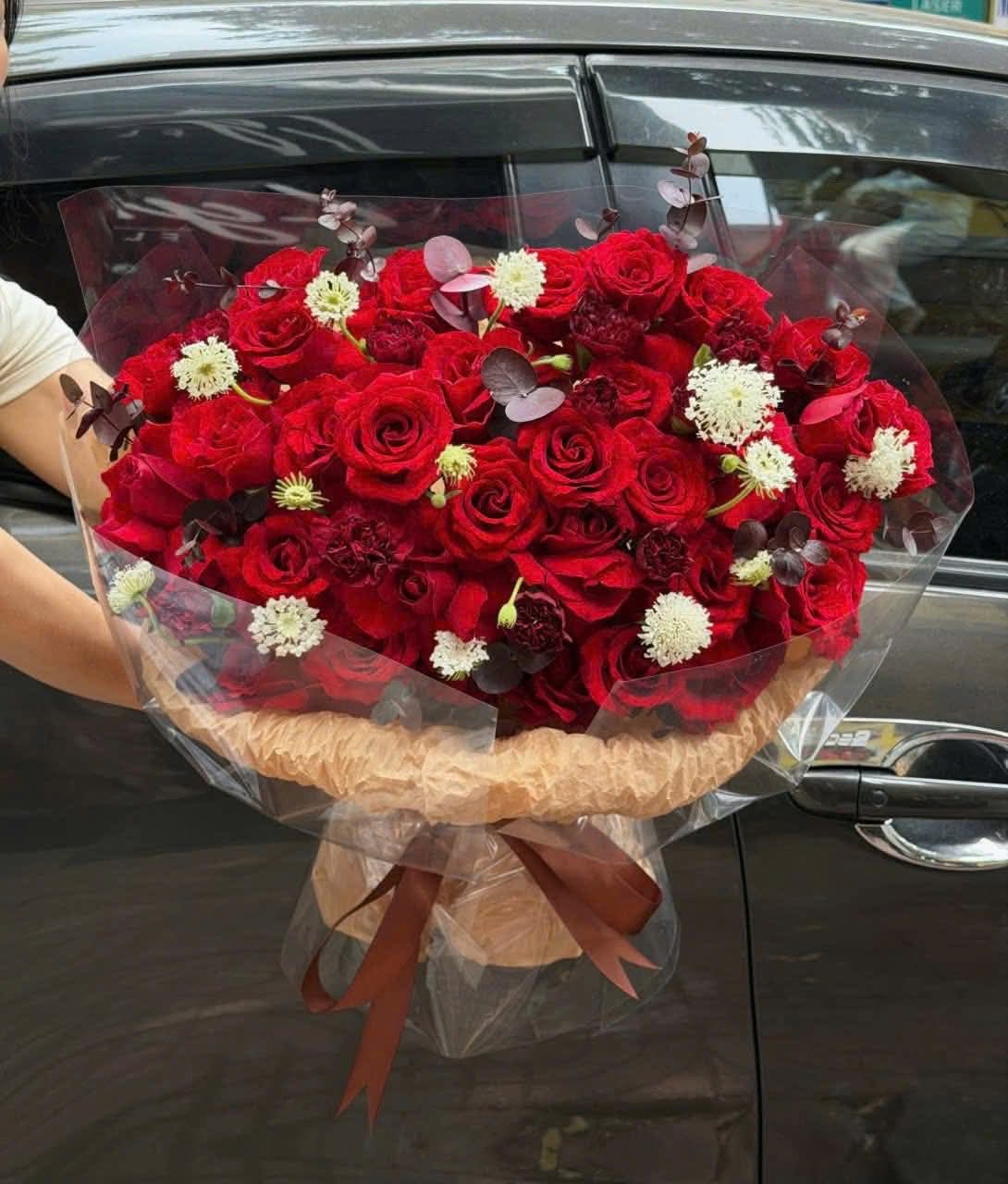 Bó hoa hồng đỏ rực rỡ sang trọng được gói cẩn thận, điểm xuyết hoa trắng nhỏ và lá phụ, thể hiện tình yêu nồng cháy. Đặt hoa