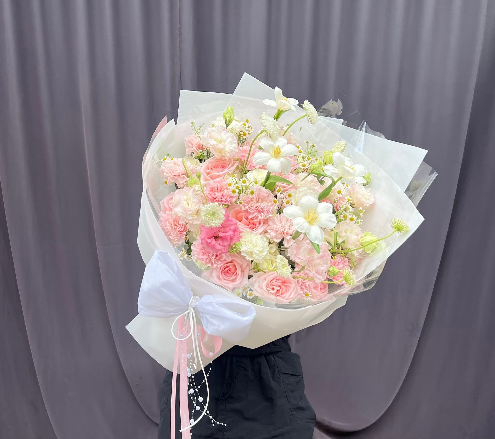 Bó hoa tươi tone hồng pastel và trắng, gói giấy sang trọng với nơ trắng lớn, trang trí ruy băng, của STflowers tại Quận 7, TP.HCM.